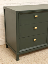 Load image into Gallery viewer, A three-drawer dresser in dark green with square gold pull handles, shown in a corner setting against a white brick wall and beige carpet.