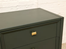 Load image into Gallery viewer, A close-up of a dark green dresser with a matte finish and a small rectangular gold pull handle, set against a white brick wall background.