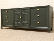 Load image into Gallery viewer, A dark green wooden sideboard with a central paneled cabinet door and four drawers on each side, accented with small gold knobs, set against a white brick wall and beige carpet.