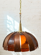 Load image into Gallery viewer, A hanging pendant light with a dome-shaped shade made of warm copper-toned metal slats and a brass-toned top cap and chain, shown against a white painted brick wall.