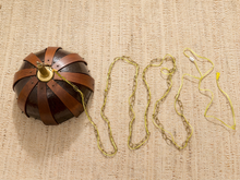 Load image into Gallery viewer, A vintage-style brass and leather ball ornament with a long chain lies on a textured beige woven fabric background, alongside a yellow cord and small yellow accessories.