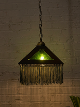 Load image into Gallery viewer, A hanging pendant lamp with a triangular dark metal shade, green glass panel, and long fringe, suspended against a white brick wall.