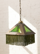 Load image into Gallery viewer, A hanging pendant light with a dark bronze metal frame, green stained glass panels, and a long fringe shade, shown against a white brick wall with sunlight and shadows.