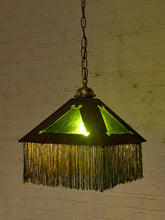 Load image into Gallery viewer, A vintage-style pendant light with a dark metal frame, green-tinted glass panels, and a long fringe hanging below, suspended by a chain against a white brick wall.