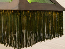 Load image into Gallery viewer, Close-up of a dark brown wooden ceiling fan with a long fringe of dark green tassels, set against a light off-white brick wall.