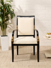 Load image into Gallery viewer, A classic high-back dining chair with a black lacquered frame, cream-colored upholstered seat and backrest, and ornate gold metal accents on the armrests, set against a white brick wall background.