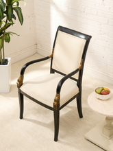 Load image into Gallery viewer, An elegant armchair with a black wood frame, cream upholstered seat and backrest, and gold decorative accents, shown in a bright room with a white brick wall, potted plant, and side table.