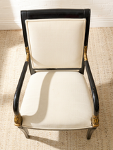 Load image into Gallery viewer, An elegant armchair with a glossy black frame, cream-colored textured fabric upholstery, and ornate gold accents on the armrests and legs, set on a woven jute rug.