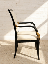 Load image into Gallery viewer, A classic high-back dining chair with a black wood frame, cream upholstered seat and back, and ornate gold gilded armrest accents, set against a white brick wall with dramatic sunlight and shadow patterns.