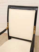 Load image into Gallery viewer, A close-up of a vintage-style armchair featuring a black lacquered frame, cream-colored textured fabric upholstery, and decorative gold-toned metal accents on the armrests, set against a white brick wall background.