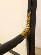 Load image into Gallery viewer, Close-up detail of a black metal frame chair with a decorative gold-toned claw-shaped corner finial and cream-colored textured sling fabric.