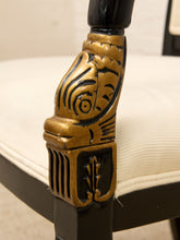 Load image into Gallery viewer, Close-up of an ornate carved wooden chair leg cap in a distressed gold finish with black detailing, attached to a black wooden frame and a cream-colored upholstered seat.