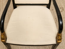 Load image into Gallery viewer, An armchair with a cream-colored ribbed fabric seat cushion and backrest, supported by a glossy black frame with ornate gold gilded arm accents, set on a beige carpet.