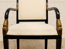 Load image into Gallery viewer, An elegant armchair with a glossy black frame, cream-colored upholstered seat and backrest, and ornate gold accents on the armrests, set against a white brick wall background.