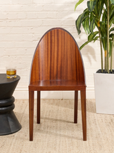 Load image into Gallery viewer, A mid-century modern wooden dining chair with a curved backrest, shown in a styled room setting with a white brick wall, a potted plant, and a small side table.