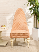 Load image into Gallery viewer, A tall upholstered accent chair with a peach and white gingham pattern, cream legs, and a high curved backrest, set in a styled room with a white brick wall, potted plant, and side table.