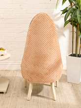 Load image into Gallery viewer, A tall, egg-shaped chair cover in a peachy beige woven pattern, shown on a wooden chair in a bright, modern room with a white brick wall and a potted plant.