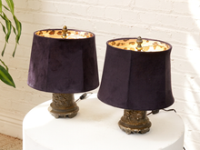 Load image into Gallery viewer, Two matching table lamps with dark plum purple velvet drum shades and distressed bronze bases, displayed on a white pedestal against a white brick wall.