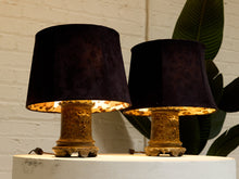 Load image into Gallery viewer, Two table lamps with dark plum velvet shades and textured gold bases, displayed on a white pedestal against a white brick wall with a green leaf in the background.