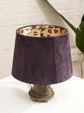 Load image into Gallery viewer, A table lamp with a dark purple velvet drum shade, a leopard-print patterned inner lining, and an ornate distressed bronze metal base, shown on a white surface against a white brick wall.