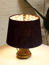 Load image into Gallery viewer, A table lamp with a dark plum purple velvet shade, a gold-toned metal finial and base, and a warm cream patterned inner shade lining, shown lit on a white surface against a white brick wall.