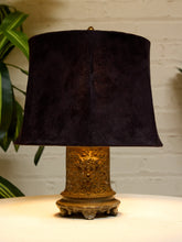 Load image into Gallery viewer, A table lamp with a dark plum purple velvet drum shade and an ornate gold-finished carved base, set on a white surface against a white brick wall with a green plant in the background.