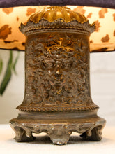 Load image into Gallery viewer, An ornate antique-style bronze metal bell or chime with a detailed face motif on the front, standing on a decorative base in front of a patterned lampshade.