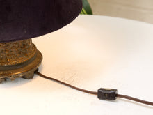 Load image into Gallery viewer, A close-up of a vintage table lamp with a dark purple velvet shade, ornate gold metal base, and a brown power cord with a black switch on a white table surface.