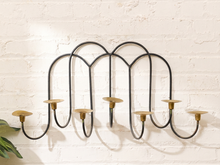 Load image into Gallery viewer, A black metal wall-mounted candelabra with five curved arms and brass candle cups, mounted on a white brick wall.