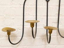 Load image into Gallery viewer, A close-up of a black metal wall-mounted candelabra with three brass candle cups against a white painted brick wall.