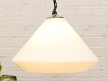 Load image into Gallery viewer, A modern pendant light with a large, tapered white glass shade suspended from a black cord against a white brick wall background.