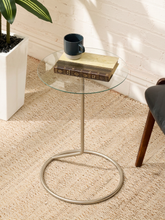 Load image into Gallery viewer, A modern round glass-top side table with a slim metal pedestal base and circular footrest, styled in a bright room with a woven rug, brick wall, plant, and books on top.