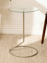 Load image into Gallery viewer, A modern side table with a clear glass round top and a curved silver metal base, set on a textured beige rug against a white brick wall.