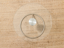 Load image into Gallery viewer, A minimalist wall-mounted circular metal hook with a brushed silver center disc, mounted on a clear circular glass or acrylic disc over a woven beige textured wall.