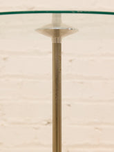Load image into Gallery viewer, A close-up of a modern table base featuring a brushed brass or bronze metal pedestal and a clear glass top, set against a light beige brick wall background.