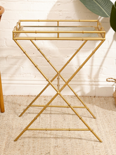 Load image into Gallery viewer, A gold-finished metal side table with a bamboo-inspired frame design and a clear glass tray top, set on a woven jute rug against a white brick wall.