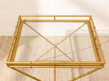 Load image into Gallery viewer, A gold-finished bamboo-style side table with a clear glass top, shown against a white brick wall and woven beige rug.
