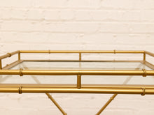 Load image into Gallery viewer, A close-up of a gold metal tray table with a glass top, featuring a bamboo-inspired frame design, set against a white brick wall background.