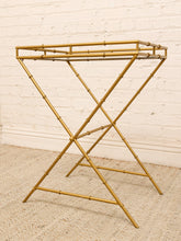Load image into Gallery viewer, A gold-finished metal stand with a bamboo-inspired jointed frame and two open top shelves, photographed against a white painted brick wall on a beige woven rug.