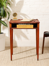 Load image into Gallery viewer, A mid-century modern wooden writing desk with tapered legs and a single drawer, styled in a room setting with a white brick wall, potted plant, and chair.