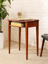 Load image into Gallery viewer, A small wooden writing desk with tapered legs and a single drawer, styled in a room setting against a white brick wall with a potted plant and a stool partially visible.