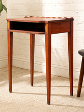 Load image into Gallery viewer, A small wooden writing desk or console table with four tapered legs, an open shelf, and a scalloped apron top, shown in a room corner against a white brick wall.