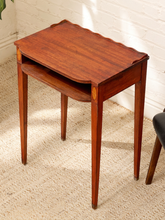 Load image into Gallery viewer, A small wooden nesting side table with a warm reddish-brown finish, featuring a scalloped top, a lower shelf, and four tapered legs, set on a textured beige rug against a white brick wall.