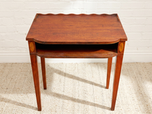 Load image into Gallery viewer, A mid-century wooden side table with a warm reddish-brown finish, featuring a scalloped apron, an open lower shelf, and oval inlay details on the front corners, set against a white brick wall and light beige rug.