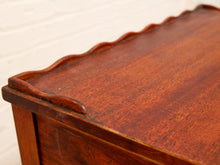 Load image into Gallery viewer, Close-up of a reddish-brown wooden chest or cabinet corner, showing the wood grain and a scalloped front edge against a white painted brick wall.