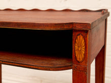 Load image into Gallery viewer, Close-up of a dark reddish-brown wooden table with a scalloped apron top edge, an open lower shelf, and a decorative sunburst inlay on the front leg, set against a white brick wall.