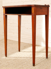 Load image into Gallery viewer, A vintage wooden writing desk or side table with four tapered legs, an open shelf, and a small drawer on the right side, shown against a white brick wall and light-colored carpet.