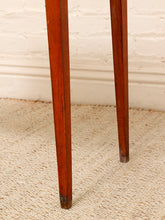 Load image into Gallery viewer, Close-up of the legs of a wooden table, showing polished reddish-brown wood grain and dark metal feet, standing on a beige woven rug against a white brick wall.