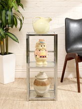 Load image into Gallery viewer, A three-tier glass and metal side table styled in a living room setting, holding a cream pitcher on top, a small ceramic character figurine in the middle, and a brown and beige lidded ceramic pot on the bottom shelf.