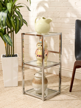 Load image into Gallery viewer, A three-tier side table with a chrome silver metal frame and clear glass shelves, styled with decorative ceramic vases in a bright room with a white brick wall, potted plant, and chair.